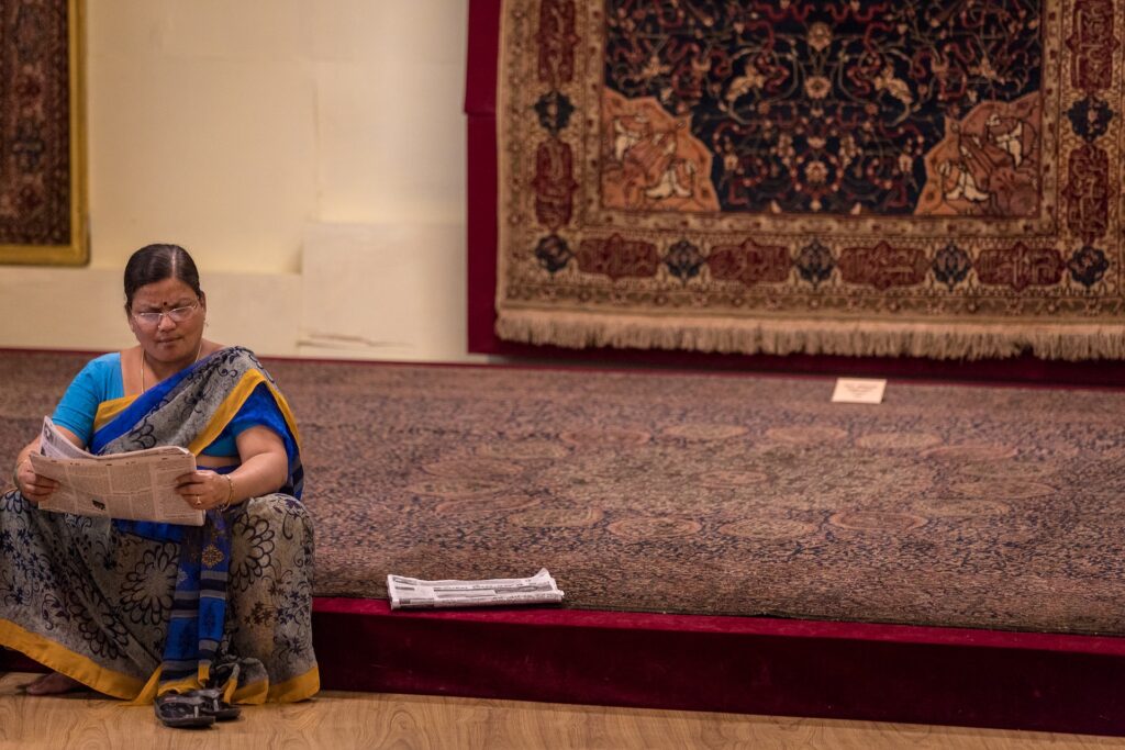 Foto de uma mulher com trajes indianos sentada sob um tapete grande, com outro tapete exposto na parede